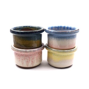 Könnte beinhalten: Vier kleine Keramiktöpfe in einem Stapel. Die Töpfe haben eine runde Form mit breitem Rand und sind in verschiedenen Farben glasiert, darunter Blau, Rosa, Gelb und Grün. Die Töpfe sind paarweise gestapelt, einer auf dem anderen.