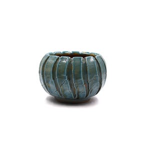 May include: A small, round ceramic bowl featuring a deep teal, glossy glaze with a distinct crackle finish. The exterior is textured with vertical, segmented ridges that wrap around the body, creating a rhythmic, ribbed appearance.