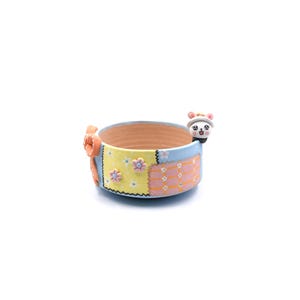 Puede incluir: Una pequeña maceta redonda de cerámica con un diseño de patchwork en amarillo, azul y rosa. Una figura de oso de dibujos animados está unida al lateral. La maceta tiene un interior marrón claro y un lazo decorativo naranja.