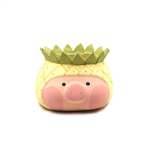 May include: A ceramic planter shaped like a pig wearing a pineapple costume. The pig has a pink face with round eyes and a snout. The pineapple top is yellow with a cross-hatched pattern and a crown of green leaves around the opening.