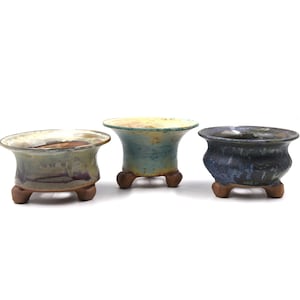 Op de afbeelding: Drie keramische bonsai potten. De potten zijn in tinten blauw, groen en bruin, met een getextureerde afwerking. Elke pot heeft een brede, uitlopende rand en staat op drie kleine, bruine voetjes.