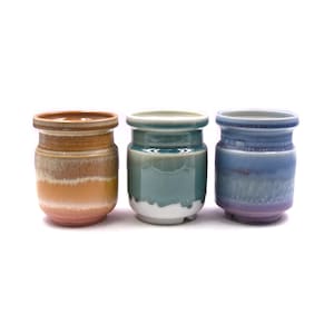 Pode incluir: Três vasos de cerâmica em diferentes cores e esmaltes. O vaso à esquerda é laranja e marrom, o do meio é verde e branco, e o da direita é roxo e azul.