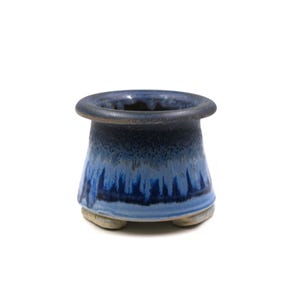 May include: A small, handcrafted ceramic pot with a dark blue rim and a gradient of blues down the sides. The base is a light tan color. The pot has a rounded shape and sits on three small feet.