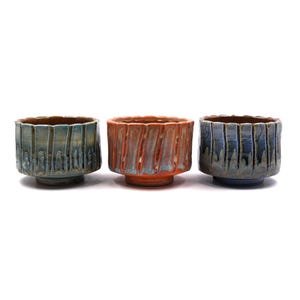 May include: Three ceramic bowls arranged in a row, each featuring a fluted, vertical ribbed exterior texture. The left bowl is blue-grey, the center bowl is a vibrant orange-red, and the right bowl is a deep blue, all with a glazed finish.
