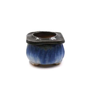 May include: A small ceramic planter with a rounded body featuring a blue and white drip glaze finish. The top rim is square-shaped with a dark charcoal grey glaze, and the base is supported by three small, short feet for stability.
