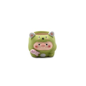 May include: A small, round ceramic planter shaped like a pig wearing a green hooded animal costume. The pig has a pink snout, rosy cheeks, and holds a small pink fish. The hood features white-tipped ears and a small black nose detail on top.