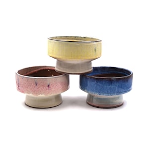 May include: Three ceramic pedestal bowls in yellow, pink, and blue. Each bowl has a wide, shallow basin and a short, rounded pedestal base. The bowls are arranged in a triangular formation against a white background, showcasing their colorful glazed surfaces.