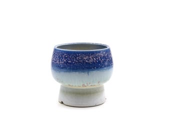 May include: A blue and white ceramic planter with a speckled glaze. The planter has a wide, round base and a slightly flared top.
