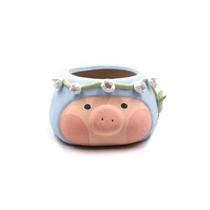 May include: A ceramic planter shaped like a pig's face, painted in light blue, pink, and beige. The planter has a floral crown and a round opening for plants. The pig has black eyes and a pink snout.