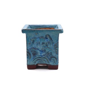 May include: A square, turquoise ceramic planter with a wave pattern. The planter has a textured surface and a dark brown base. The interior is a contrasting orange-brown color. This decorative pot is suitable for small plants.