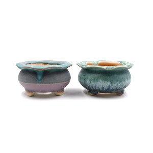 May include: Two ceramic flower pots with a scalloped rim and three feet. The pot on the left is a light purple color with a teal glaze. The pot on the right is a teal color with a green glaze.