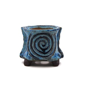Może przedstawiać: Mała, kwadratowa ceramiczna doniczka z niebieskim szkliwem i spiralnym wzorem. Doniczka ma trzy małe nóżki i teksturowaną powierzchnię. Wnętrze ma kontrastujący brązowy kolor. Dekoracyjny element do małych roślin lub sukulentów.