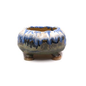 May include: A small ceramic bowl with three short legs, featuring a rounded shape and a wide opening. The exterior has a reactive glaze with a cream-colored top section that transitions into a deep blue drip pattern over a muted olive green base.