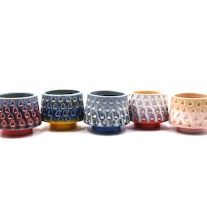 Puede incluir: Cinco tazas de cerámica dispuestas en fila, cada una con un exterior texturizado con hendiduras repetitivas en forma de gota. Las tazas muestran un degradado de colores que incluye rojo, azul, amarillo, blanco y rosa sobre una base circular.