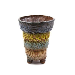 May include: A handcrafted ceramic planter with a textured, layered design. The planter features a dark blue rim, a yellow and green midsection, and a blue and brown base. It stands on three small feet, showcasing a unique, artistic aesthetic.