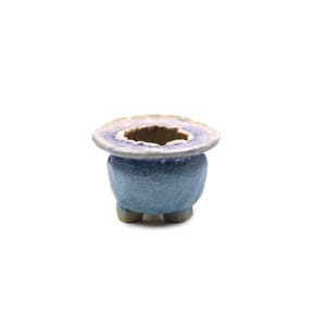 May include: A small, round ceramic planter with a textured, light blue glazed exterior. The vessel features a wide, flat, light purple rim with a rough, uneven edge and is supported by three short, sturdy feet at the base, creating a tripod design.