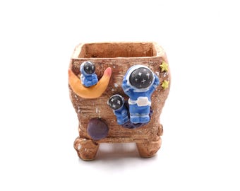 May include: A small, square, brown ceramic planter with a space theme. The planter features blue astronaut figures, a crescent moon, stars, and a purple planet. The planter has a textured surface and is elevated on small feet.