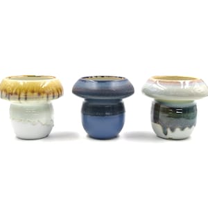 May include: Three ceramic mushroom-shaped planters in different colors. The planter on the left is white with a brown top, the middle planter is blue, and the planter on the right is white with a green and blue top.