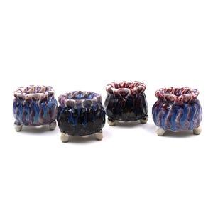 May include: Four small, ceramic pots with a cauldron shape. The pots are glazed in shades of blue, purple, and black, with a textured, dripped effect. Each pot has three small, white feet and a ruffled rim.