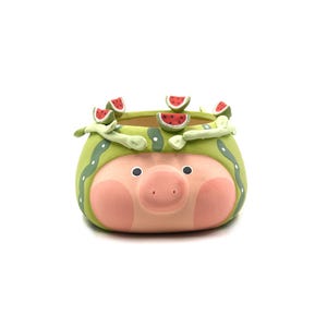 May include: A ceramic planter shaped like a pig wearing a green watermelon rind hat with small watermelon slice decorations. The pig has a light pink face, dark eyes, and a snout, while the green hat features white stripes and dots.