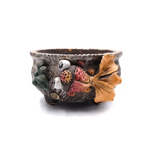 May include: A brown ceramic planter with a goldfish design. The planter has a textured surface and is decorated with green, red, orange, and white paint. The goldfish has large eyes and a long tail.