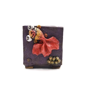 May include: Purple ceramic planter with a 3D goldfish design. The fish has red, orange, and yellow scales and large white eyes. The planter has a textured surface and a small base.