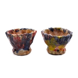 Puede incluir: Dos pequeñas macetas de cerámica hechas a mano con un aspecto rugoso y texturizado. Una maceta presenta pintura roja, azul y blanca, mientras que la otra tiene amarilla, azul y roja. Ambas tienen una forma orgánica única y son adecuadas para plantas pequeñas.
