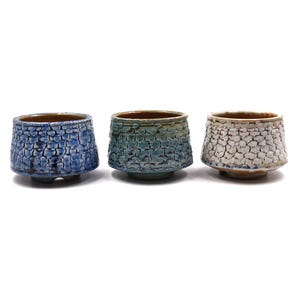 May include: Three ceramic cups with a textured, crackled glaze finish. The first cup is deep blue, the second is a gradient of teal and blue, and the third is white with brown undertones. Each cup features a wide rim and a small, raised base.