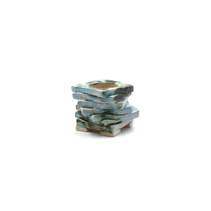 May include: A stacked ceramic candle holder with a square top and base. The holder is composed of layered, irregularly shaped pieces in shades of blue, green, and tan. The design has a rustic, handcrafted appearance.