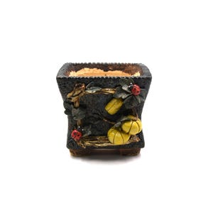 May include: A square, dark gray ceramic planter with a textured surface. The planter is decorated with yellow gourds, green leaves, and red ladybugs. The rim of the planter has a textured, light brown appearance.