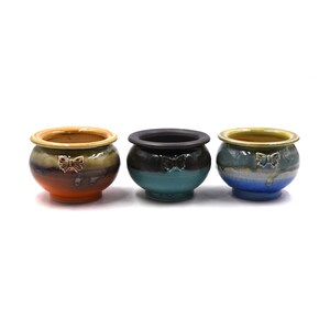 May include: Three small ceramic pots with a rounded shape and a wide rim. Each pot features a small butterfly emblem on the front. The left pot is orange and brown, the center pot is black and teal, and the right pot is blue and grey.