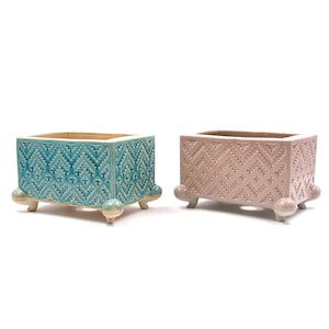 May include: Two ceramic planters with geometric patterns. The planter on the left is teal blue with a white pattern. The planter on the right is pink with a white pattern. Both planters have four small feet.