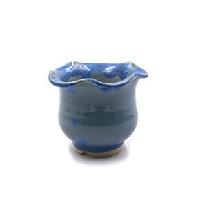 May include: A small, handmade ceramic vase with a wavy rim. The vase is a muted blue-gray color with darker blue accents around the rim and base. The interior is a lighter blue. The vase has a rounded body and a slightly flared opening.