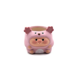 May include: A pink ceramic planter shaped like a pig. The planter has a round opening and is painted with a pig face, including a snout, eyes, and ears. The planter is designed to hold small plants or succulents.