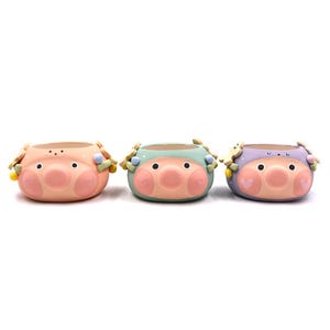 Può includere: Tre vasi in ceramica a forma di muso di maiale nei colori pesca, verde menta e lavanda. Ogni vaso presenta un muso prominente, due occhi neri, guance rosa con contorni a cuore e piccoli accenti floreali decorativi sui lati.