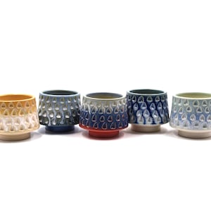 May include: Five ceramic cups arranged in a row, each featuring a textured exterior with a repeating teardrop pattern. The cups display a gradient of colors including yellow, blue, red, and light green, all set against a plain white base.