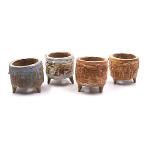 May include: Four small, handmade ceramic pots with a textured, rustic appearance. Each pot is cylindrical with three small legs. The pots are glazed in shades of blue, white, brown, and yellow, creating a unique, earthy aesthetic.
