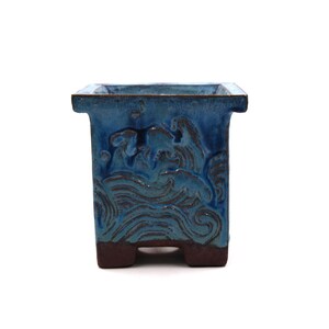Może przedstawiać: Kwadratowa ceramiczna donica z teksturowanym wzorem fal oceanu. Donica jest w żywym niebieskim kolorze z brązowymi akcentami i ciemnobrązową podstawą. Pojemnik ma unikalny, artystyczny wygląd, odpowiedni dla małych roślin.