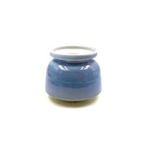 May include: A small, round ceramic container with a blue glaze. The container has a wide, open top with a white rim and a slightly flared base. The blue color graduates from a darker shade at the base to a lighter shade near the top.