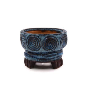 Puede incluir: Una pequeña maceta de cerámica azul con un diseño en espiral en la parte superior. La maceta tiene un interior marrón oscuro y se apoya en tres pequeños pies marrones oscuros. El exterior tiene un acabado texturizado y esmaltado.