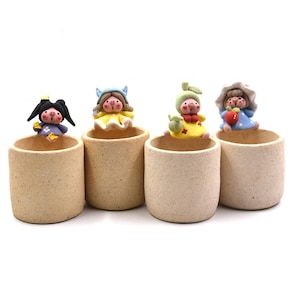 May include: Four small, beige ceramic pots each topped with a whimsical figurine. The figurines have painted faces and wear colorful outfits. One holds a star, another a flower, a third a small bag, and the last a red apple.