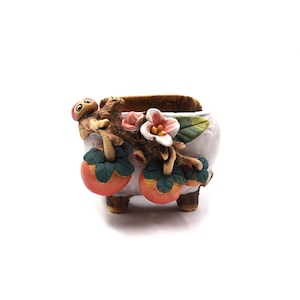 Puede incluir: Una pequeña maceta de cerámica blanca con un diseño decorativo. La maceta presenta una rama esculpida con flores rosas, hojas verdes y fruta naranja. La maceta está sostenida por tres pequeños pies.