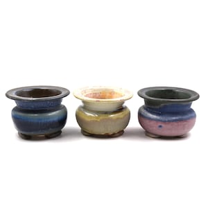 May include: Three small, ceramic pots with a rounded shape and a wide, dark rim. The pots are glazed in shades of blue, green, and tan, with a speckled or mottled appearance. Each pot has three small feet.