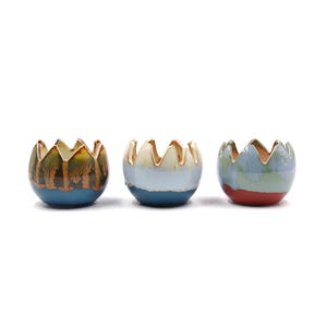 Puede incluir: Tres macetas de cerámica con forma de huevo y bordes festoneados. Las macetas están esmaltadas en diferentes colores: azul, verde y rojo.