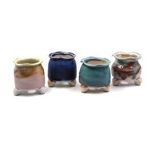 May include: Four small ceramic pots in shades of pink, brown, deep blue, teal, and maroon. Each pot has a ruffled rim and rests on three decorative feet. These pots are suitable for small plants or succulents.