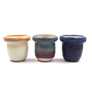 May include: Three small ceramic pots in different colors. The pot on the left is white with a light brown rim. The middle pot is a dark blue with a light pink rim. The pot on the right is a dark blue with a light blue rim.