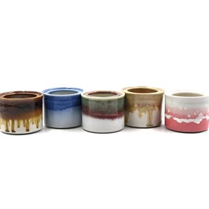 May include: Five cylindrical ceramic pots arranged in a row. The first is brown with a white drip glaze, the second is blue, the third is green and white with a red band, the fourth is tan with a drip glaze, and the fifth is pink and white.