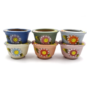 Op de afbeelding: Set van drie keramische bloempotten met geschilderde bloemenmotieven. Elke pot heeft een bijpassende schotel. De potten zijn blauw, groen en grijs met gele en oranje bloemen.