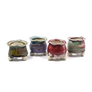 May include: Four small, handcrafted ceramic pots in shades of green, purple, red, and green-grey. Each pot has a scalloped rim and sits on three small, rounded feet. The pots are glazed with a glossy finish.