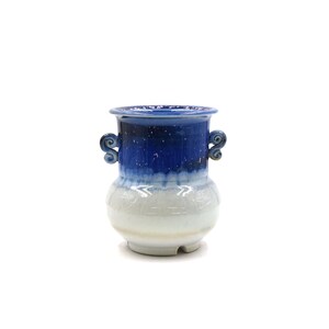 Puede incluir: Un jarrón de cerámica azul y blanco con dos asas. El jarrón tiene un esmalte azul moteado y un esmalte blanco en la mitad inferior.